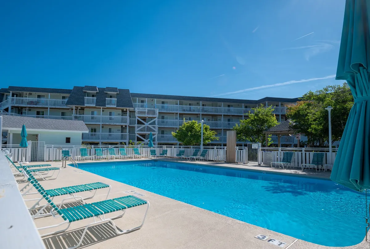 An outdoor pool surrounded by cheerful lounge chairs, with the backdrop of a residential building and lush green trees. The scene is lively yet relaxed, suggesting family-friendly enjoyment and summertime gatherings.