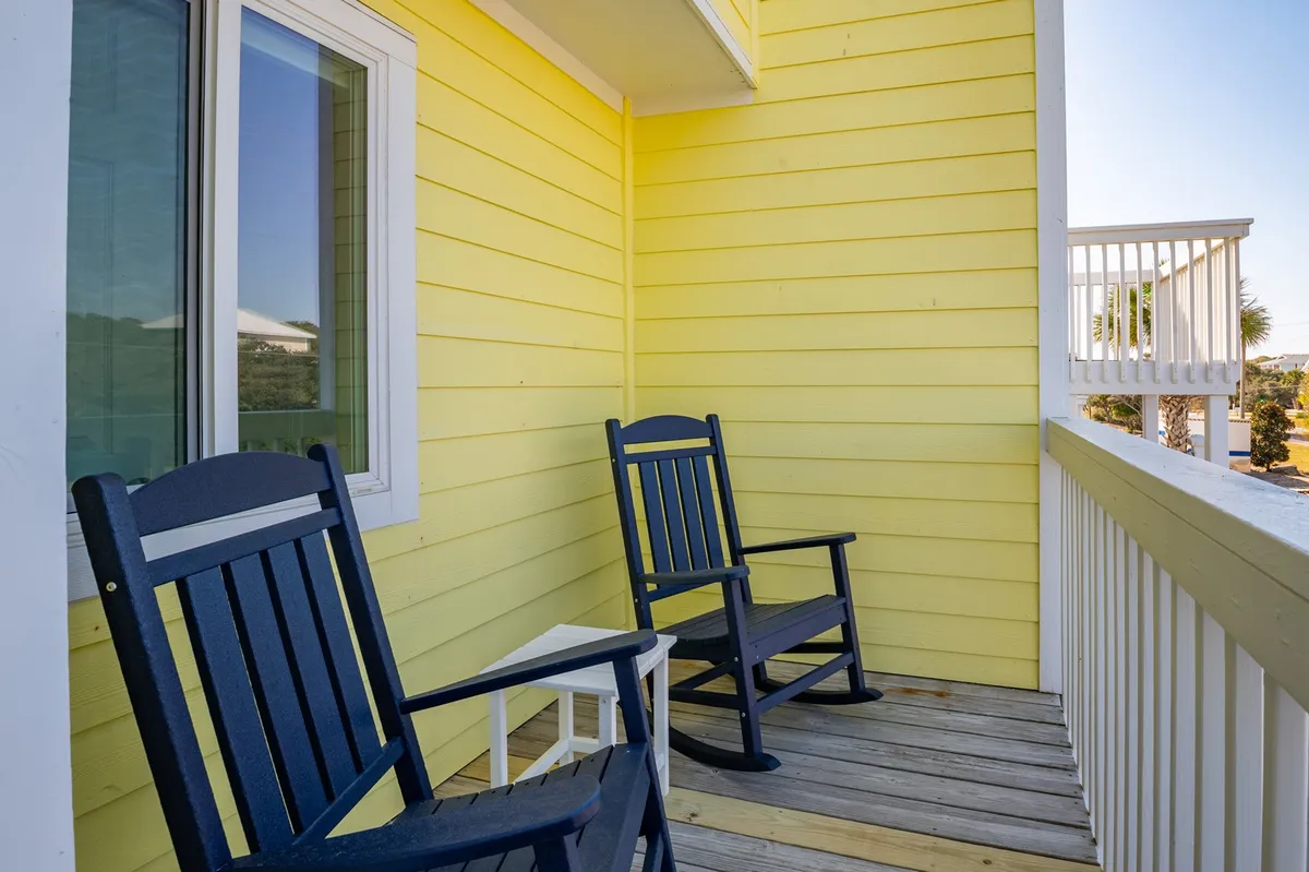 A welcoming oceanfront porch with classic white railings, yellow walls, and inviting rocking chairs. The view beyond hints at the ocean, creating an ideal setting for sipping coffee at sunrise or enjoying evening breezes while lounging outside.