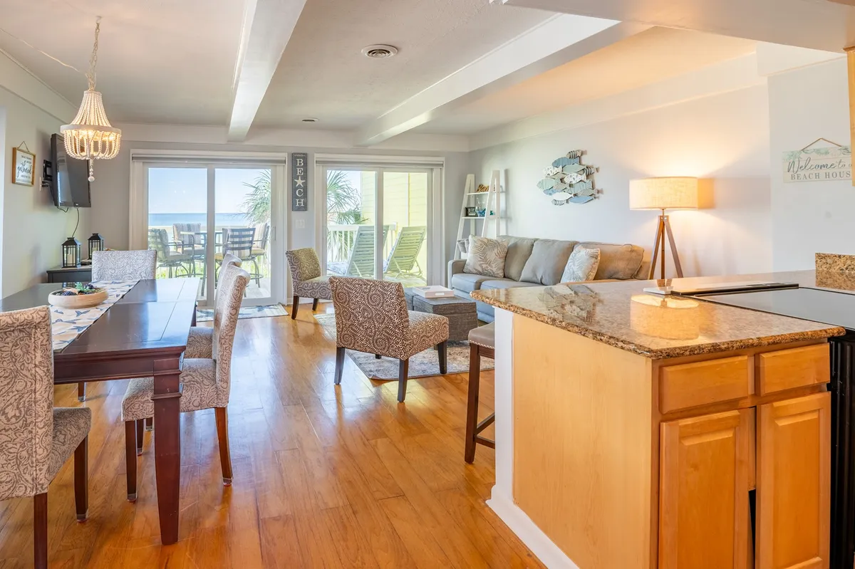 A spacious, sunlit open-concept living area that seamlessly connects the living room, dining table, and kitchen. Large windows and sliding glass doors lead out to an inviting outdoor deck, allowing natural light to flood the space. The living room features comfortable seating and the dining area has a large table, perfect for meals or game nights. The kitchen is visible in the background, supporting easy conversation and togetherness.