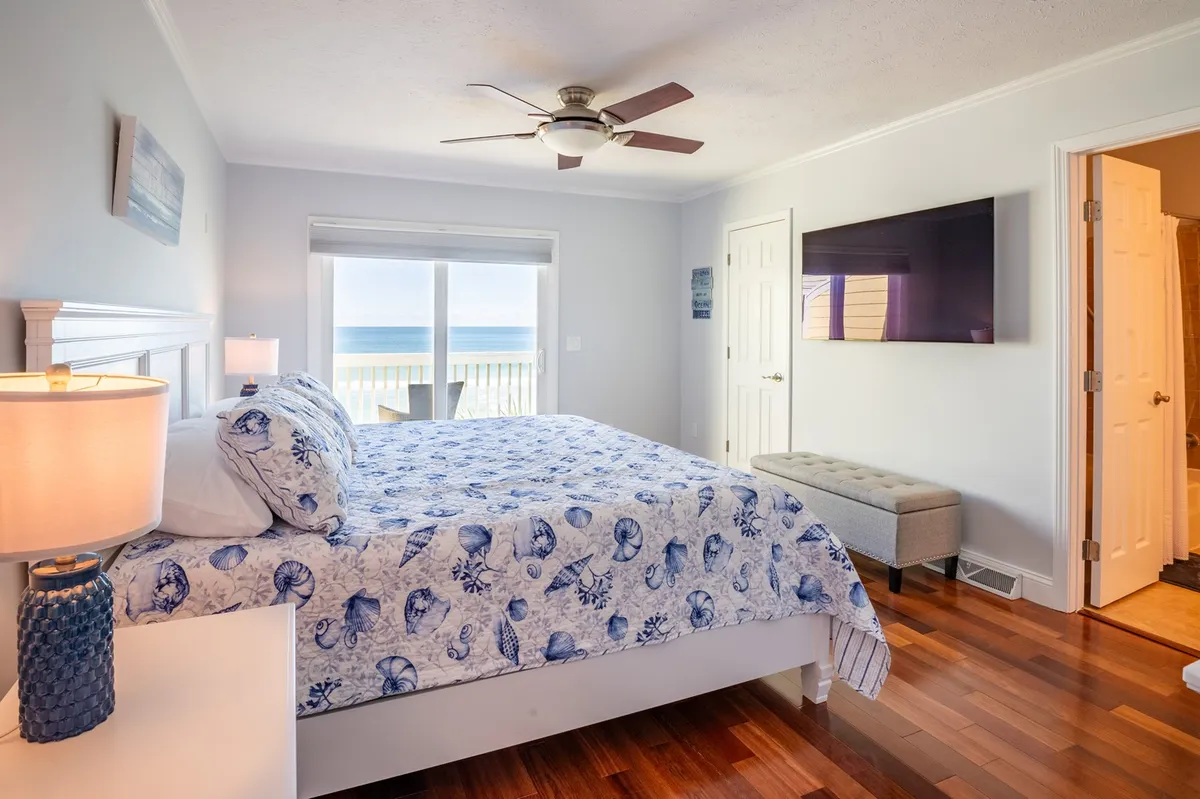 This tranquil bedroom features a king-sized bed with ocean-themed bedding, a large window framing stunning ocean views, a ceiling fan overhead, and a wall-mounted TV. Soft natural light fills the space, enhancing the calming ambiance.