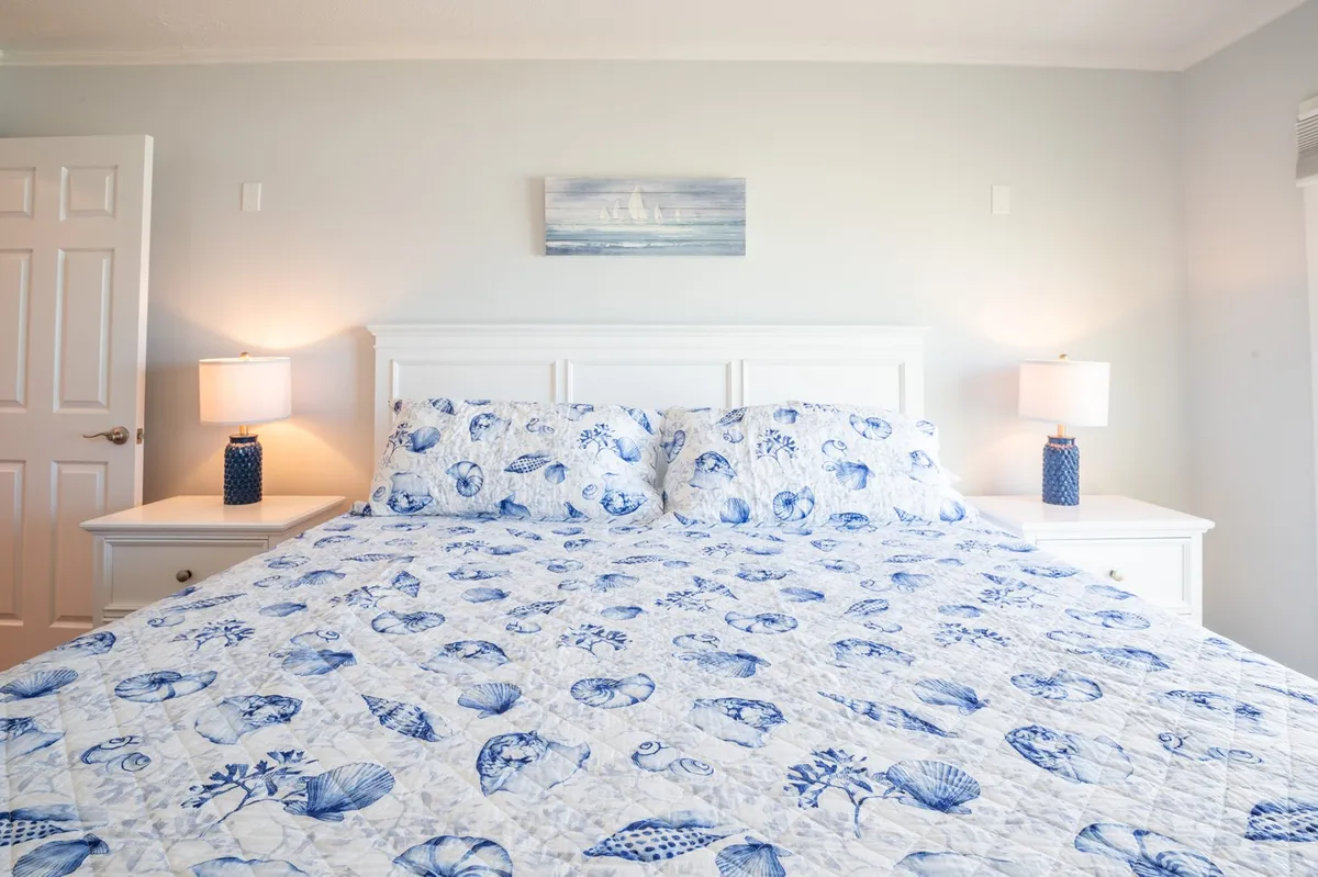 A tranquil bedroom scene with a bed featuring a patterned quilt, blue lamps on each side, nautical-themed artwork above, and soft daylight streaming in. The ambiance is peaceful and inviting.
