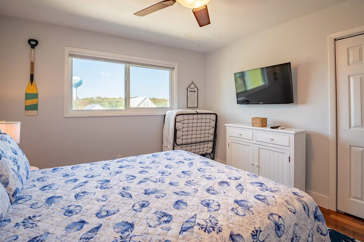 A cozy bedroom with a blue and white quilted bed, a window inviting in soft daylight, a wall-mounted TV, and a decorative paddle accenting the wall. The room feels intimate yet bright, perfect for guest comfort.