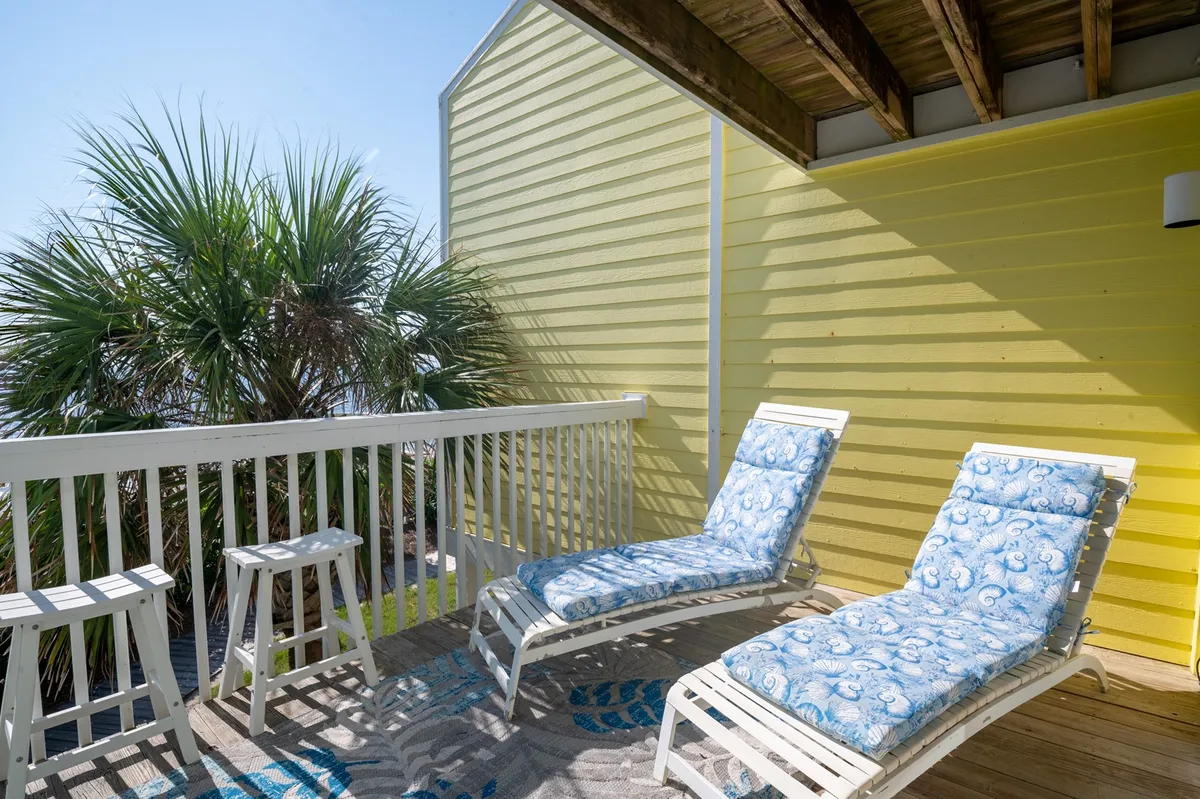 An inviting exterior lounge area with comfortable lounge chairs, a palm tree for shade, and a vibrant yellow wall, evoking a sense of relaxation and leisure. The setting suggests a tranquil space for reading, unwinding, or enjoying the scenic view—possibly of the ocean.