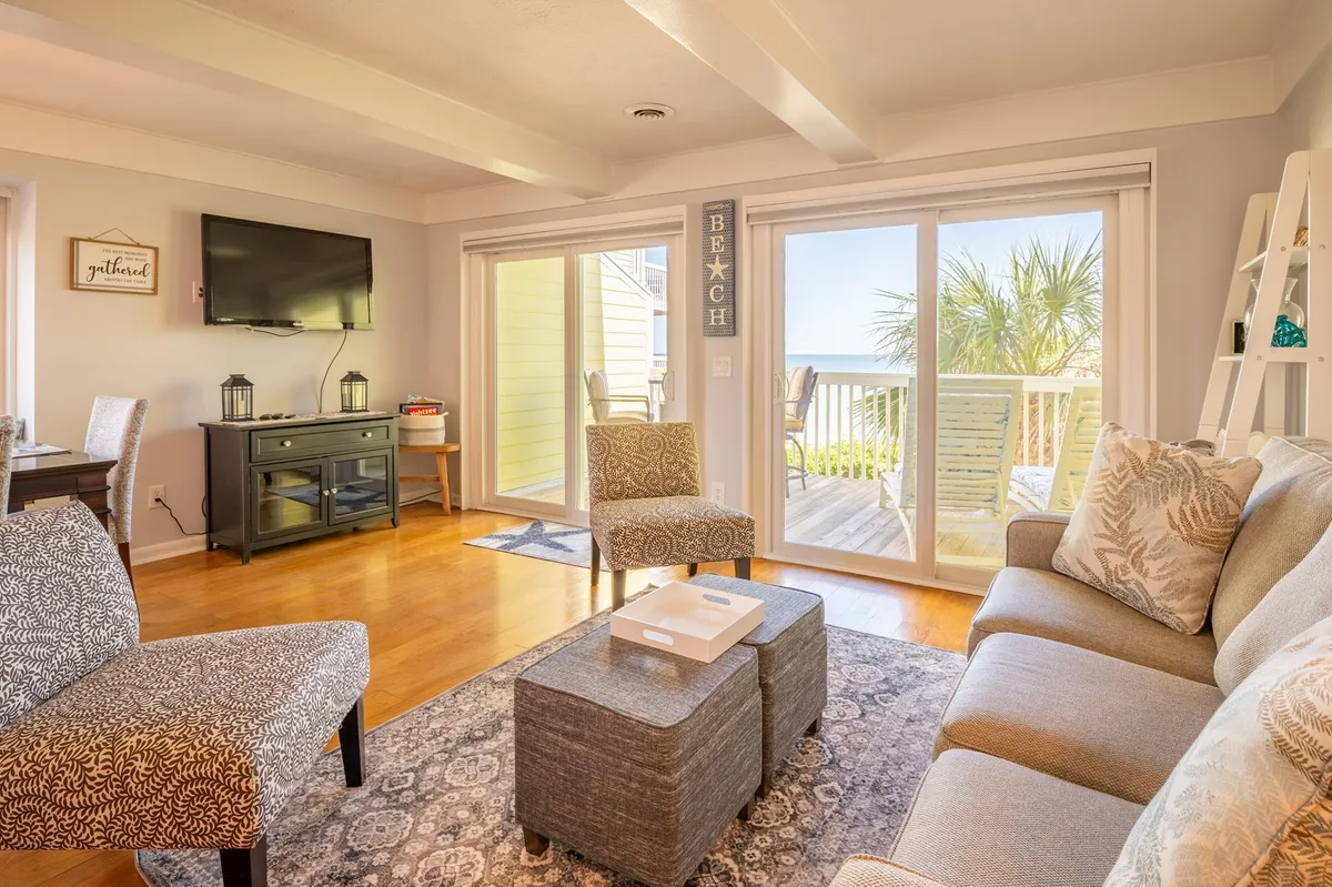 A cozy living room with a plush sofa, coffee table, and large windows that offer a glimpse of the ocean beyond. There's a 65-inch smart TV mounted for movie nights and a decorative sign that adds a coastal touch. The natural light creates an inviting atmosphere, ideal for relaxing or socializing.