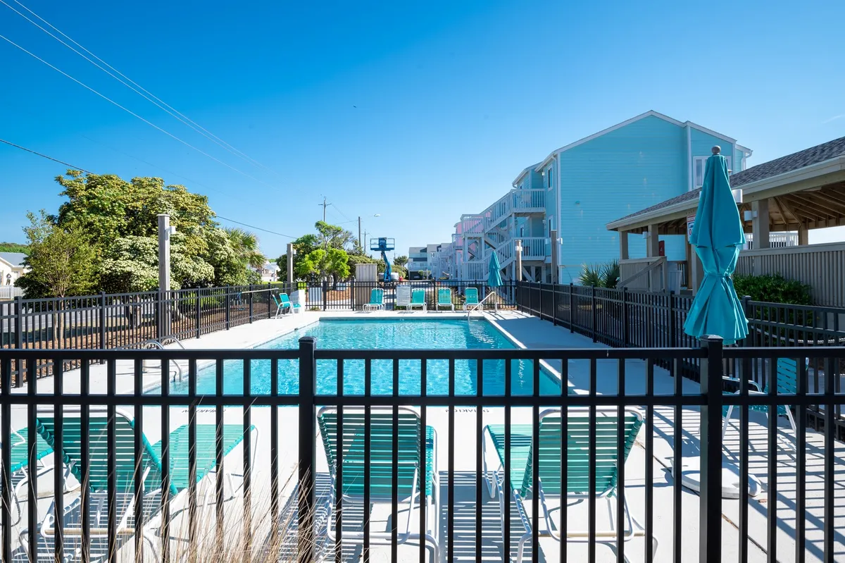 A sparkling outdoor swimming pool surrounded by lounge chairs and umbrellas, with a perimeter fence and lush greenery in the background. The pool area looks well-maintained and ready for leisurely swims, sunbathing, or socializing by the water.