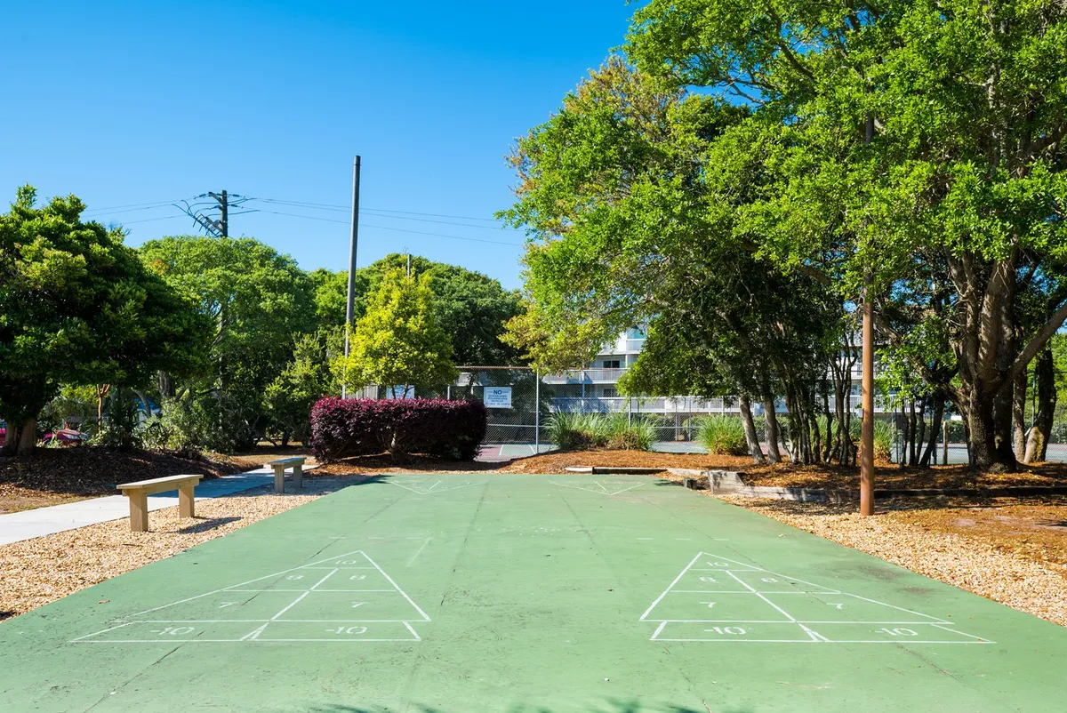 A shuffleboard court set among leafy trees, with benches nearby for relaxing and watching the fun. The setting is open and playful, encouraging outdoor games and social time for all ages.