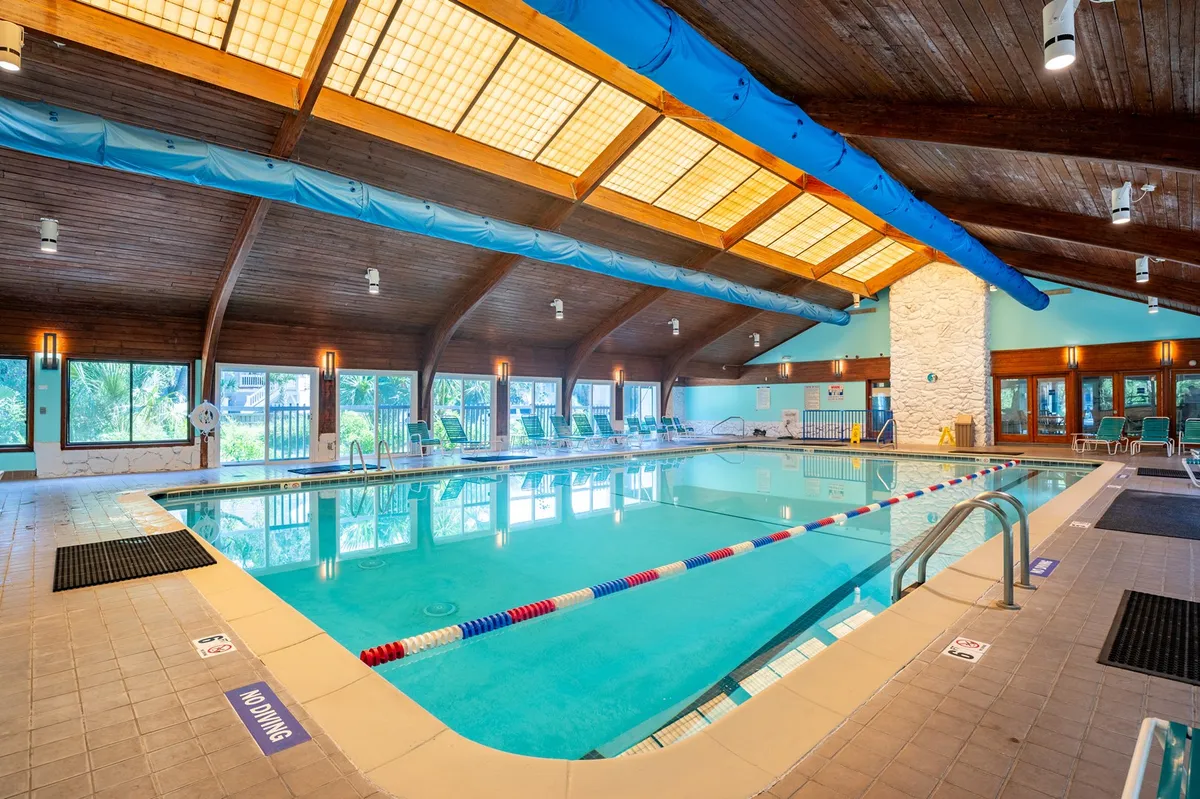A spacious indoor swimming pool area with striking wooden beams, large windows flooding the room with natural light, and comfortable lounge chairs lining the pool. The atmosphere is tranquil and inviting, and the space is designed for both swimming and social gatherings.