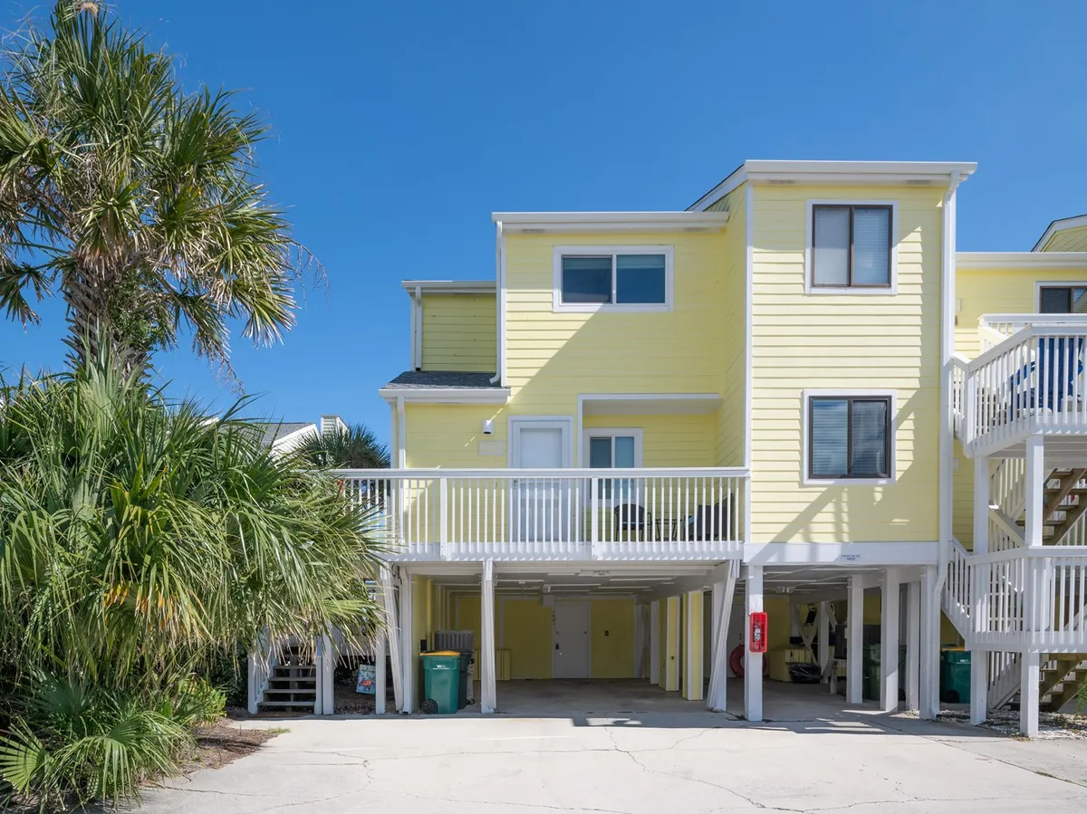 An inviting exterior scene featuring the yellow house, white railings on the deck, and lush palm trees. The deck area suggests a perfect spot for morning coffee or evening relaxation while soaking in the sea breeze.