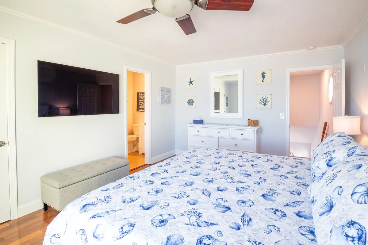 A bedroom with seashell-patterned bedding, a large wall-mounted TV, and a dresser with decorative items. Sunlight filters in, creating a calm and inviting space ideal for relaxation and entertainment.