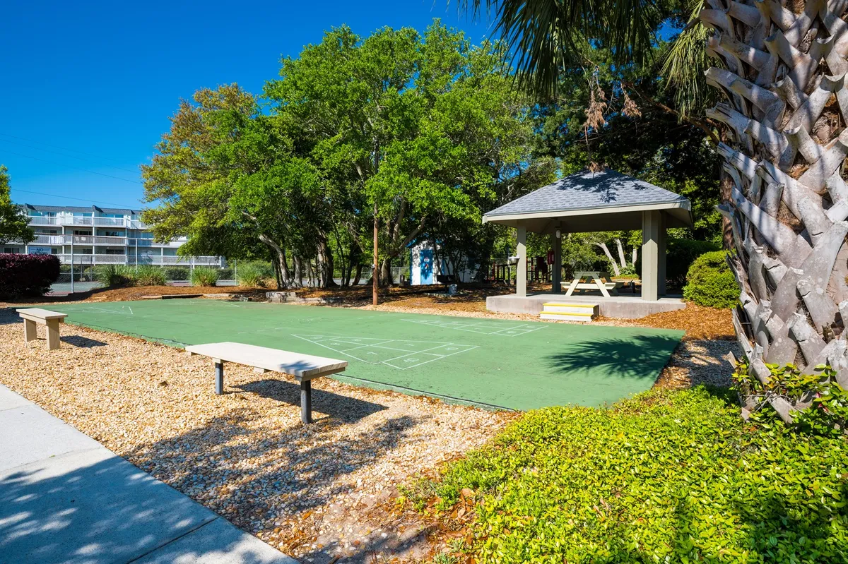 A lush green outdoor play and activity area with a gazebo, surrounding trees, and benches. The space is ideal for outdoor games, shuffleboard, and family relaxation, capturing a sense of playful community spirit.